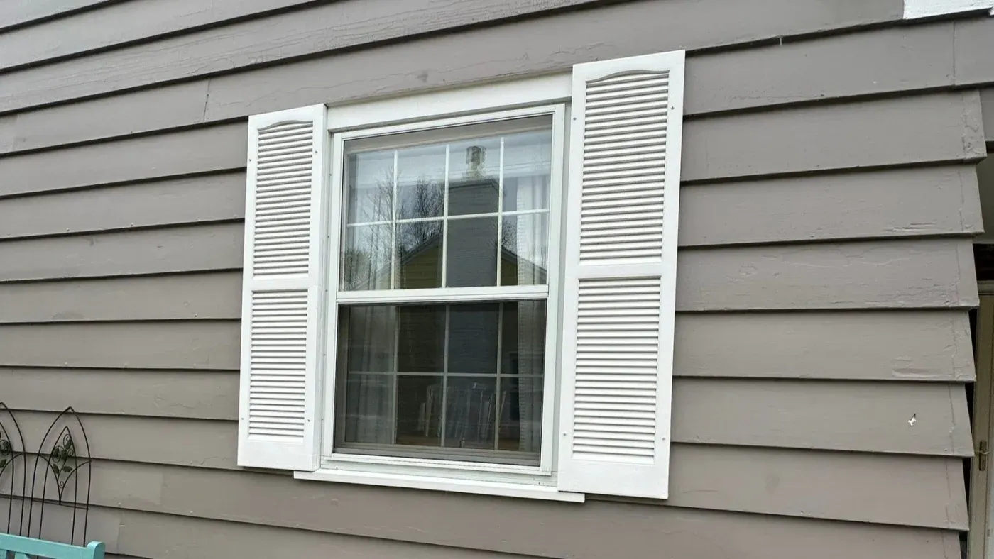 Double-hung window with white louver shutters on gray horizontal siding, reflecting bare trees and sky
