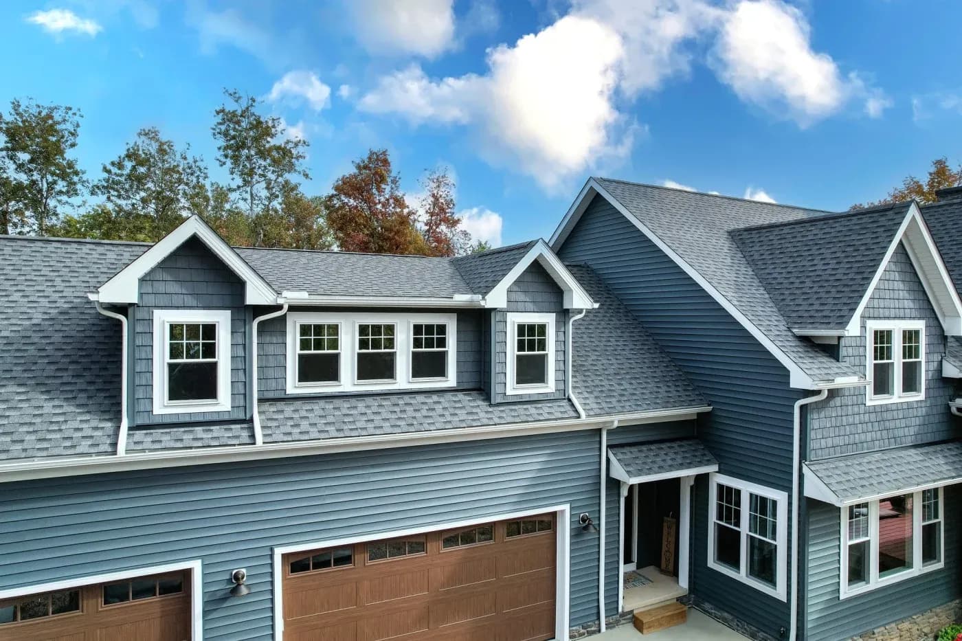 Modern residential home with blue-gray siding, white trim, multiple gabled rooflines with gray shingles, white-framed windows, and brown garage doors, set against autumn foliage and clear sky