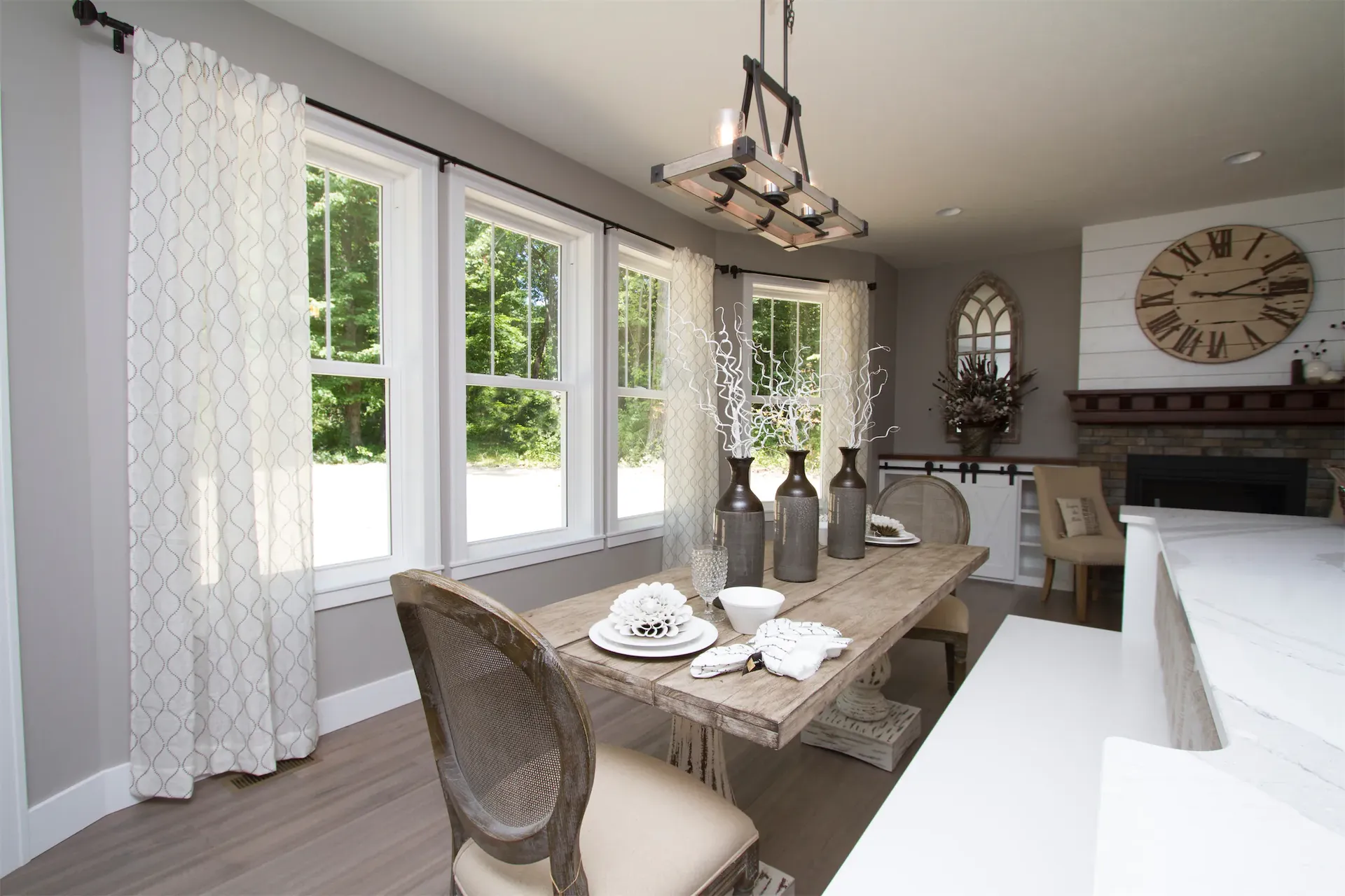 Interior view of a rustic modern dining room featuring large double-hung white grid windows with patterned drapes, a rustic wooden table set with decorative vases, and a stone fireplace in the background.